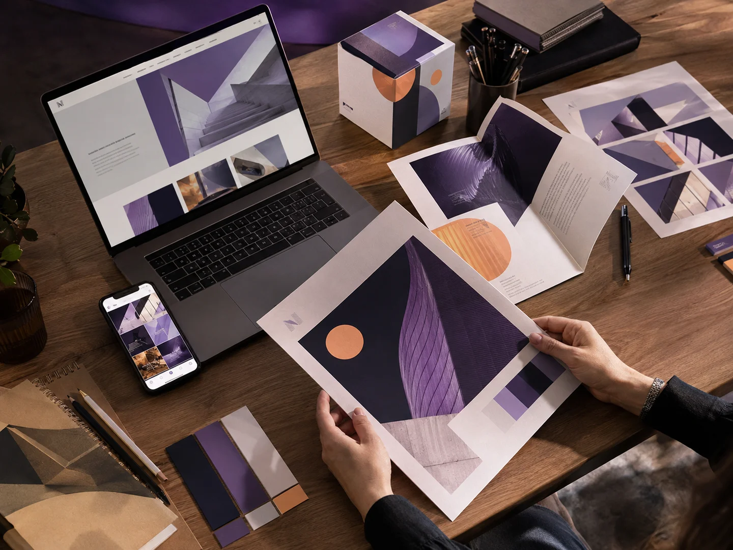 Coordinated violet and apricot campaign across a website, phone, brochure, posters, and packaging on a designer’s desk.